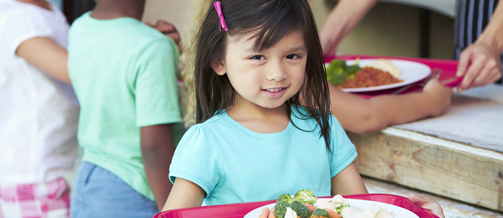 Ett litet barn håller i en bricka med mat i skolmatsalen. 